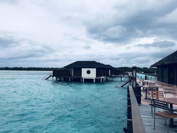 A tranquil scene featuring overwater villas on stilts, set against a backdrop of a cloudy sky. The water is a vibrant turquoise, and there are several wooden tables and chairs on a nearby deck, suggesting a place for relaxation.