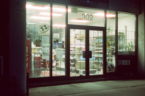 A pharmacy is seen through a glass storefront at night, brightly lit from inside. The doors and windows are transparent, revealing shelves stocked with various products. Signs are displayed on the glass, and there's a Coca-Cola vending machine visible. The interior lights highlight the organized arrangement of goods on the shelves.