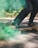 A person wearing black pants and black sneakers rides a skateboard on a paved surface. Vibrant green smoke surrounds the skateboard, creating a dynamic and energetic atmosphere. The background features lush greenery, suggesting an outdoor setting.