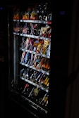 A vending machine filled with various snacks and beverages, including chips, chocolate bars, and canned drinks. The machine is dimly lit, highlighting the items inside through the clear glass panel.