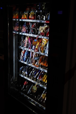 A vending machine filled with various snacks and beverages, including chips, chocolate bars, and canned drinks. The machine is dimly lit, highlighting the items inside through the clear glass panel.