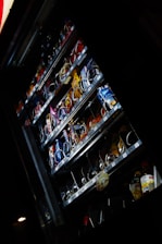 A vending machine containing various packaged snacks and drinks is partially illuminated in a dark environment. The machine's shelves display a variety of candy bars, chips, cookies, and bottled beverages. Some rows of products appear almost empty, indicating that they have been frequently purchased.