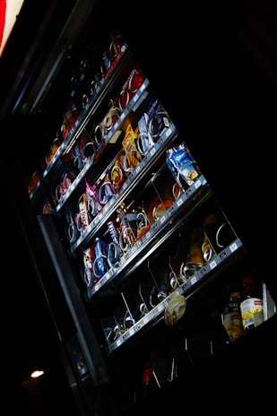 A vending machine containing various packaged snacks and drinks is partially illuminated in a dark environment. The machine's shelves display a variety of candy bars, chips, cookies, and bottled beverages. Some rows of products appear almost empty, indicating that they have been frequently purchased.