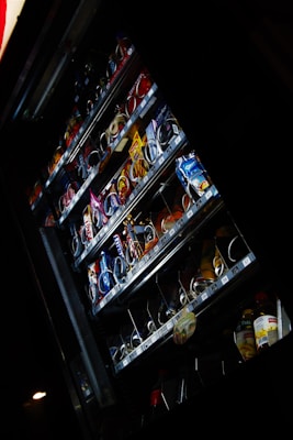 A vending machine containing various packaged snacks and drinks is partially illuminated in a dark environment. The machine's shelves display a variety of candy bars, chips, cookies, and bottled beverages. Some rows of products appear almost empty, indicating that they have been frequently purchased.