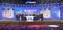 A group of people dressed in formal attire stands on a stage with a Nasdaq podium in the center. The backdrop displays the word 'BLUECITY' along with various smaller images of individuals. Confetti is falling, creating a celebratory atmosphere.