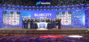 A group of people dressed in formal attire stands on a stage with a Nasdaq podium in the center. The backdrop displays the word 'BLUECITY' along with various smaller images of individuals. Confetti is falling, creating a celebratory atmosphere.