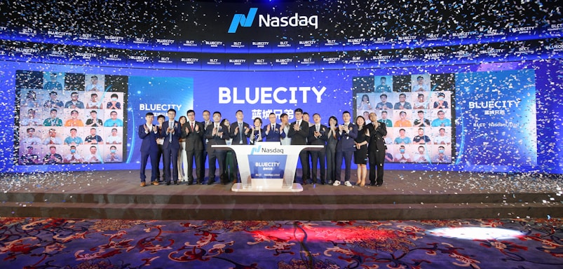 A group of people dressed in formal attire stands on a stage with a Nasdaq podium in the center. The backdrop displays the word 'BLUECITY' along with various smaller images of individuals. Confetti is falling, creating a celebratory atmosphere.