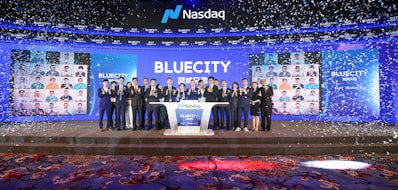 A group of people dressed in formal attire stands on a stage with a Nasdaq podium in the center. The backdrop displays the word 'BLUECITY' along with various smaller images of individuals. Confetti is falling, creating a celebratory atmosphere.