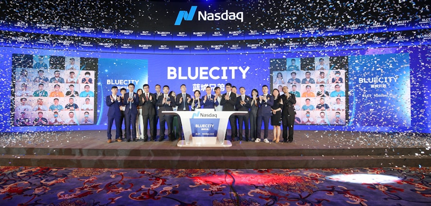 A group of people dressed in formal attire stands on a stage with a Nasdaq podium in the center. The backdrop displays the word 'BLUECITY' along with various smaller images of individuals. Confetti is falling, creating a celebratory atmosphere.