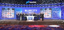 A group of people dressed in formal attire stands on a stage with a Nasdaq podium in the center. The backdrop displays the word 'BLUECITY' along with various smaller images of individuals. Confetti is falling, creating a celebratory atmosphere.