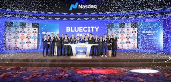 A group of people dressed in formal attire stands on a stage with a Nasdaq podium in the center. The backdrop displays the word 'BLUECITY' along with various smaller images of individuals. Confetti is falling, creating a celebratory atmosphere.
