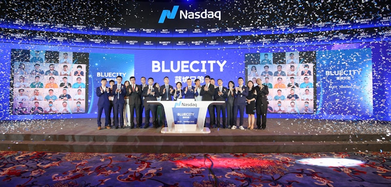 A group of people dressed in formal attire stands on a stage with a Nasdaq podium in the center. The backdrop displays the word 'BLUECITY' along with various smaller images of individuals. Confetti is falling, creating a celebratory atmosphere.