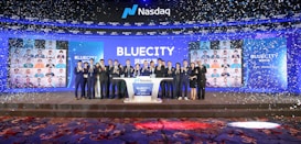 A group of people dressed in formal attire stands on a stage with a Nasdaq podium in the center. The backdrop displays the word 'BLUECITY' along with various smaller images of individuals. Confetti is falling, creating a celebratory atmosphere.