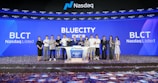 A group of people standing on a stage during a Nasdaq listing ceremony. The background features large digital screens displaying 'Bluecity' and ticker symbols such as 'BLCT'. Confetti is scattered on the floor, indicating a celebratory event.
