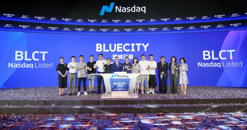 A group of people standing on a stage during a Nasdaq listing ceremony. The background features large digital screens displaying 'Bluecity' and ticker symbols such as 'BLCT'. Confetti is scattered on the floor, indicating a celebratory event.