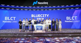 A group of people standing on a stage during a Nasdaq listing ceremony. The background features large digital screens displaying 'Bluecity' and ticker symbols such as 'BLCT'. Confetti is scattered on the floor, indicating a celebratory event.