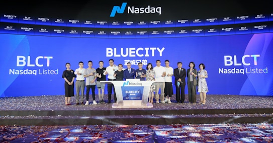 A group of people standing on a stage during a Nasdaq listing ceremony. The background features large digital screens displaying 'Bluecity' and ticker symbols such as 'BLCT'. Confetti is scattered on the floor, indicating a celebratory event.