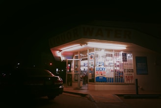A dimly lit convenience store exterior at night with bright fluorescent lights inside illuminating the interior. The store has bars on the windows and signs advertising lottery retail, discounts, and other products. A car is parked in the shadowy foreground.