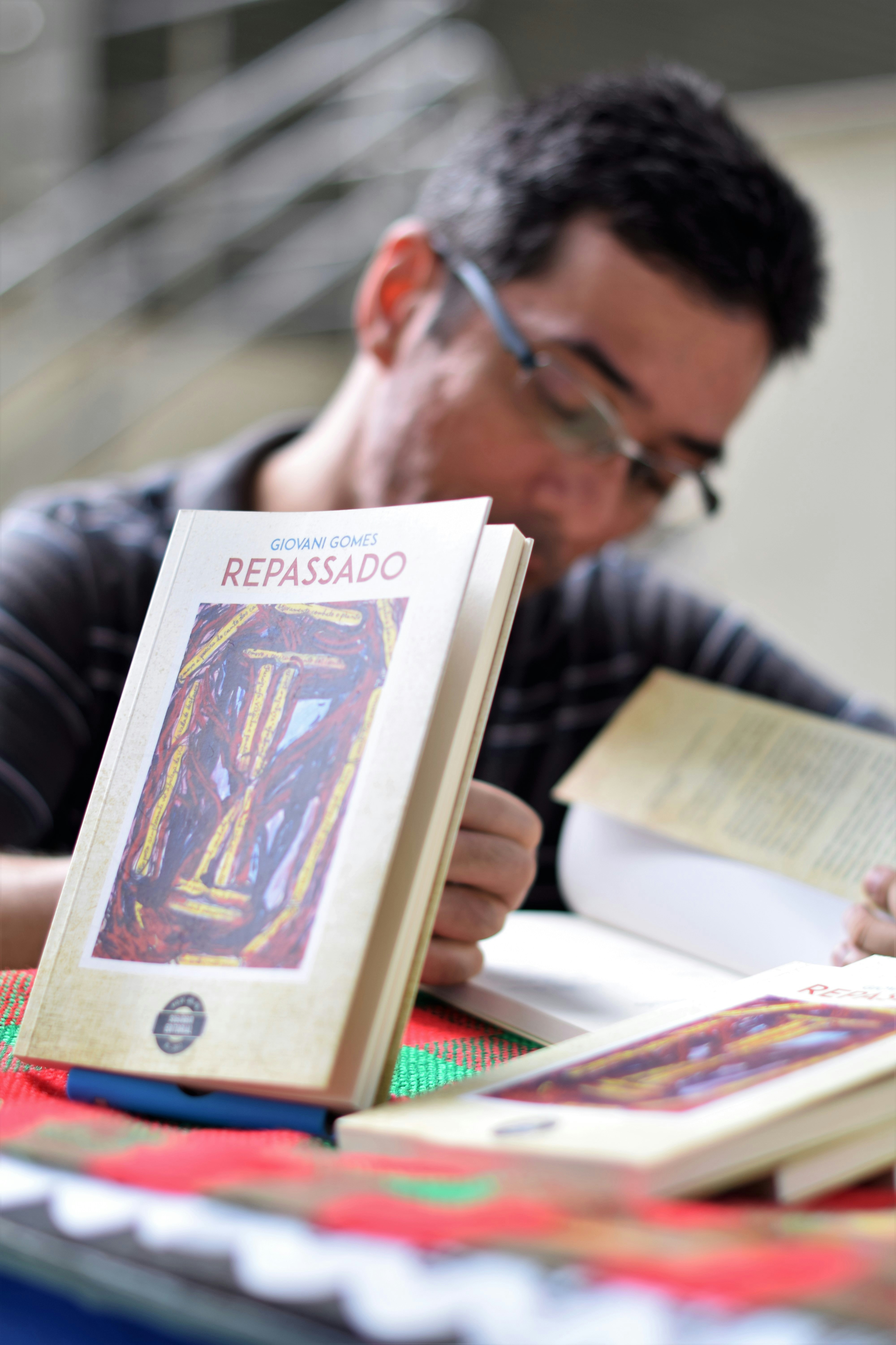 A person is sitting at a table, engaged with a book. The focus is on several books with a cover design featuring abstract art and the title 'Repassado'. The person appears to be reading, creating a scholarly or contemplative atmosphere.