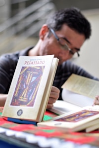 A person is sitting at a table, engaged with a book. The focus is on several books with a cover design featuring abstract art and the title 'Repassado'. The person appears to be reading, creating a scholarly or contemplative atmosphere.
