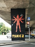 A street art mural under a concrete overpass depicts a surreal figure with multiple arms and a triangular head, featuring a single eye on its torso. The word 'PANICZ' is boldly painted below in yellow letters. A traffic light and pedestrian crossing sign are visible nearby, with some greenery and parked cars in the background.