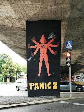 A street art mural under a concrete overpass depicts a surreal figure with multiple arms and a triangular head, featuring a single eye on its torso. The word 'PANICZ' is boldly painted below in yellow letters. A traffic light and pedestrian crossing sign are visible nearby, with some greenery and parked cars in the background.
