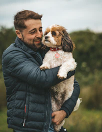 man in black leather jacket holding white and brown long coated small dog
