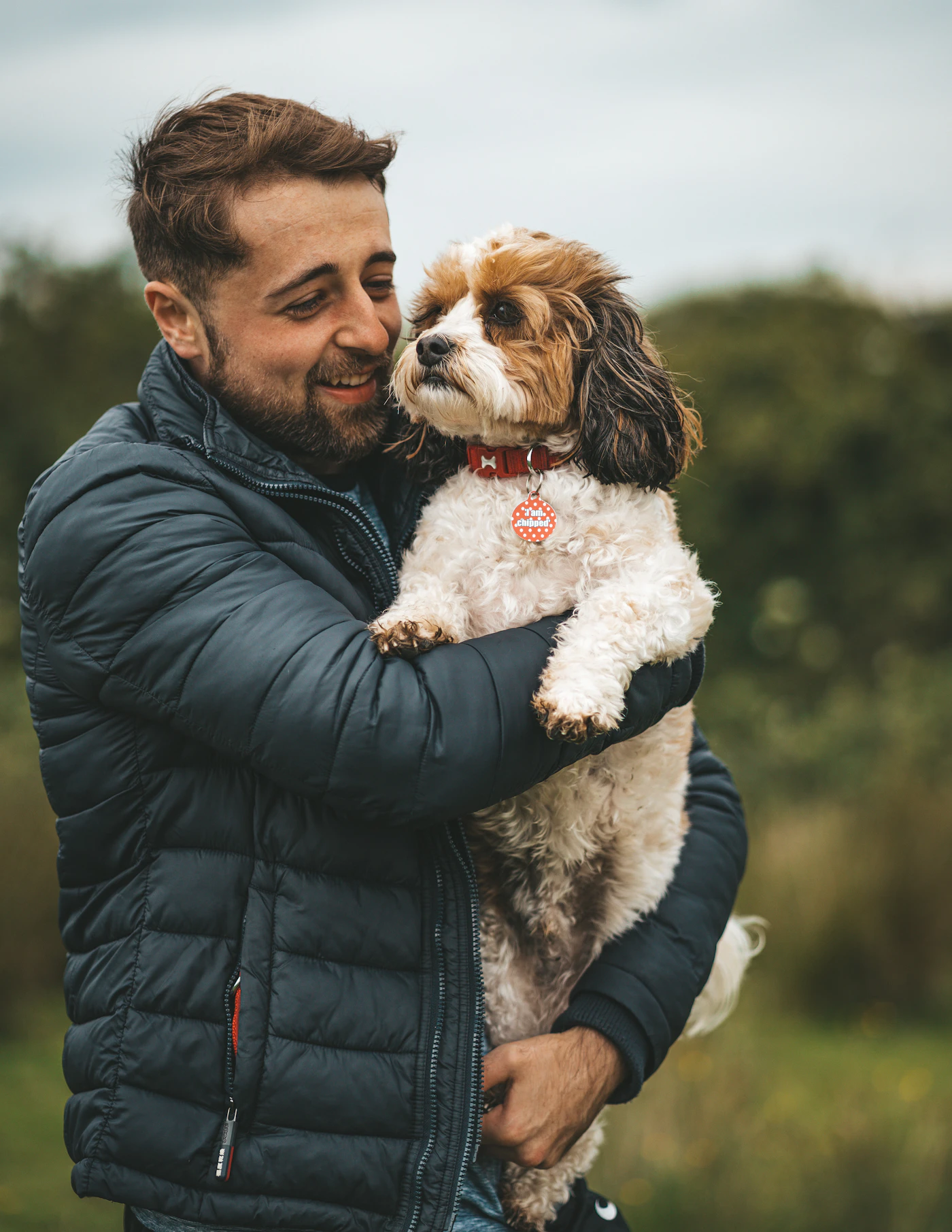 A smiling man holding his dog close against his cheek outdoors in an open green field