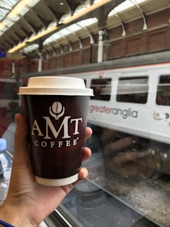 A person holds an AMT Coffee cup in a train station, with a 'greater anglia' train visible through the window. The setting suggests travel and a casual moment of having coffee while waiting or traveling.