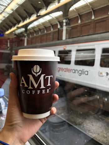 A person holds an AMT Coffee cup in a train station, with a 'greater anglia' train visible through the window. The setting suggests travel and a casual moment of having coffee while waiting or traveling.