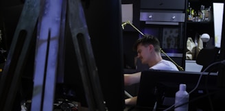 A person is seated at a desk in a dimly lit room that appears to be an art or creative studio. There is a large easel in the foreground, partially blocking the view. Shelves in the background hold bottles, boxes, and artwork. The person seems focused and engaged in an activity, such as drawing or writing.