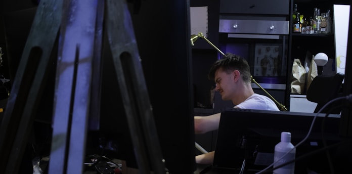A person is seated at a desk in a dimly lit room that appears to be an art or creative studio. There is a large easel in the foreground, partially blocking the view. Shelves in the background hold bottles, boxes, and artwork. The person seems focused and engaged in an activity, such as drawing or writing.