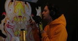 A person wearing an orange hoodie is speaking into a microphone while holding a smartphone. There is a colorful, abstract mural with symmetrical designs in the background. The person is wearing large headphones and seems to be in a recording or audio setup.