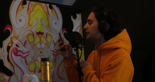 A person wearing an orange hoodie is speaking into a microphone while holding a smartphone. There is a colorful, abstract mural with symmetrical designs in the background. The person is wearing large headphones and seems to be in a recording or audio setup.