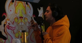 A person wearing an orange hoodie is speaking into a microphone while holding a smartphone. There is a colorful, abstract mural with symmetrical designs in the background. The person is wearing large headphones and seems to be in a recording or audio setup.