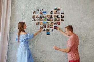 Two individuals, a woman and a man, are arranging photographs on a wall in the shape of a heart. The woman, in a blue polka-dot dress, is adding a photo while the man in a pink shirt is also engaged in placing a picture. The background is a textured, pale green wall.