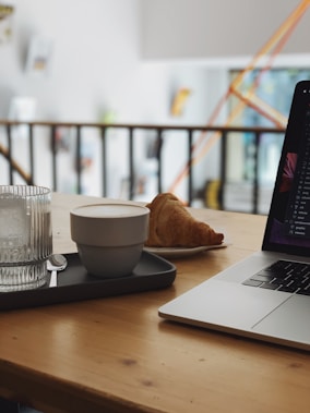 A cozy workspace setup with a warm wooden table featuring a laptop, a glass of water, a tray with a coffee cup, and a croissant. The background shows a blurred indoor setting with artwork and decorations on the wall.