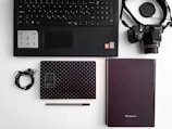A flat lay of office and tech items arranged on a white surface. Includes a black keyboard, a camera with a strap, two notebooks—one black with white dots and the other plain, a pair of earphones, and a black pen. The layout is organized and minimalist.