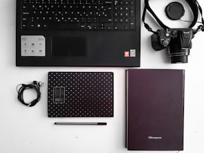 A flat lay of office and tech items arranged on a white surface. Includes a black keyboard, a camera with a strap, two notebooks—one black with white dots and the other plain, a pair of earphones, and a black pen. The layout is organized and minimalist.