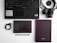 A flat lay of office and tech items arranged on a white surface. Includes a black keyboard, a camera with a strap, two notebooks—one black with white dots and the other plain, a pair of earphones, and a black pen. The layout is organized and minimalist.