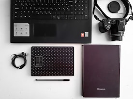 A flat lay of office and tech items arranged on a white surface. Includes a black keyboard, a camera with a strap, two notebooks—one black with white dots and the other plain, a pair of earphones, and a black pen. The layout is organized and minimalist.