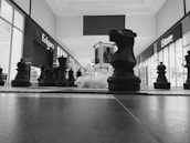 A large chess set is arranged on a shopping mall floor, with black and white pieces prominently displayed. The scene is captured from a low angle, and stores such as Edgars and iStore are visible in the background. The focus is on the knight piece at the center.
