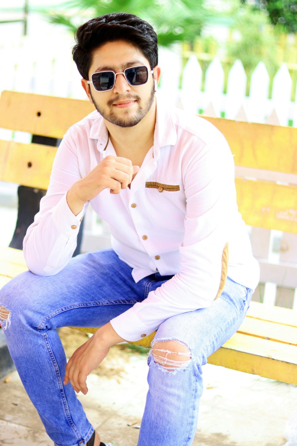 man in white dress shirt and blue denim jeans wearing black sunglasses sitting on brown wooden
