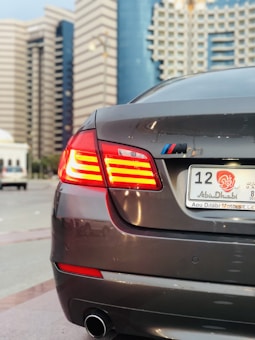 A sleek gray car with red tail lights in focus, displaying a UAE Abu Dhabi license plate in a city with modern, high-rise buildings in the background.