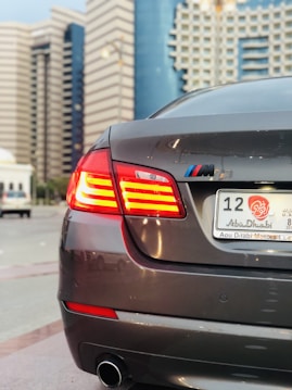 A sleek gray car with red tail lights in focus, displaying a UAE Abu Dhabi license plate in a city with modern, high-rise buildings in the background.
