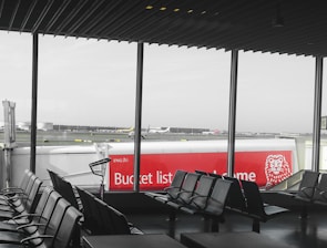 The interior of an airport departure lounge with rows of empty black chairs facing large windows. Outside, an airplane is parked near a walkway connected to the terminal. A red advertisement with the words 'Bucket list' and a lion logo on a jet bridge is prominently visible. The sky appears overcast, and the entire scene seems calm and quiet.