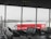 The interior of an airport departure lounge with rows of empty black chairs facing large windows. Outside, an airplane is parked near a walkway connected to the terminal. A red advertisement with the words 'Bucket list' and a lion logo on a jet bridge is prominently visible. The sky appears overcast, and the entire scene seems calm and quiet.