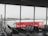 The interior of an airport departure lounge with rows of empty black chairs facing large windows. Outside, an airplane is parked near a walkway connected to the terminal. A red advertisement with the words 'Bucket list' and a lion logo on a jet bridge is prominently visible. The sky appears overcast, and the entire scene seems calm and quiet.