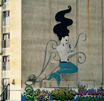 A large, artistic mural on a concrete wall depicts a stylized mermaid with flowing black hair and blue tail, holding a ship in her hands. Surrounding the mural at the base are colorful graffiti tags and street art featuring various abstract designs and shapes. The wall itself displays a worn, textured surface.