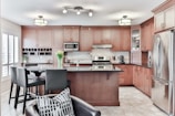 A modern kitchen with wooden cabinetry and stainless steel appliances features a central island with a black countertop. Above the island hang contemporary light fixtures. A vase with tulips sits on the island, adding a touch of color. The kitchen also has a wine rack, a built-in microwave, and a gas stove.